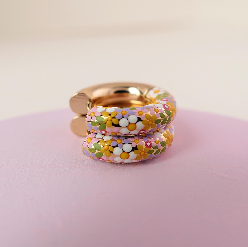Puede incluir: Un par de pendientes de aro dorados con un dise&ntilde;o floral. Los pendientes est&aacute;n cubiertos con un patr&oacute;n colorido de flores rosas, amarillas, blancas y verdes.