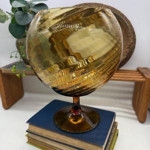 May include: A large, amber-colored glass with a round bowl and a short stem. The glass has a textured, swirling design. The glass is sitting on top of a stack of blue books. Decorative glassware.