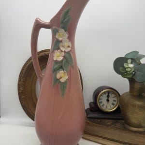 Puede incluir: Una jarra de cerámica alta de color rosa polvoriento con un cuello esbelto y alargado y una sola asa. El frente de la jarra está decorado con un relieve moldeado de flores blancas con centros amarillos y hojas verdes a lo largo del cuerpo.