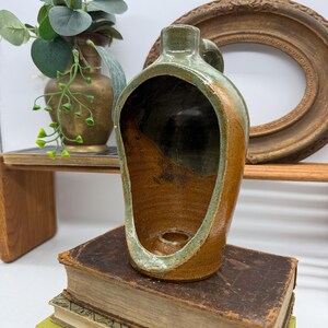 Könnte beinhalten: Eine handgefertigte Keramikvase mit einem einzigartigen, offenen Design. Die Vase hat eine Mischung aus braunen, grünen und beigen Farben, mit einem kleinen Griff und einer kreisförmigen Öffnung am Boden. Die Vase steht auf einem Stapel alter Bücher.