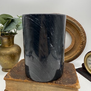 May include: A cylindrical black marble container with white veining. The container is sitting on a vintage book. A gold-colored vase with greenery and a decorative gold-colored frame are in the background.