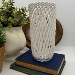 Puede incluir: Un jarrón de cerámica blanca tejida con un diseño de enrejado. El jarrón cilíndrico mide aproximadamente 25 cm de alto y se encuentra sobre una pila de libros. El fondo incluye un jarrón blanco con vegetación y un espejo dorado.