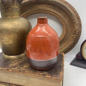 Könnte beinhalten: Eine kleine, abgerundete Keramikvase mit einer glänzenden orangefarbenen Glasur im oberen Bereich und einem matten, dunkelbraunen Band am Boden. Die Vase hat einen schmalen, kurzen Hals und eine einfache, kreisförmige Öffnung an der Oberseite.