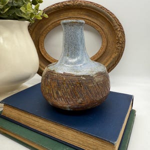 Puede incluir: Un jarrón de cerámica hecho a mano con una parte inferior marrón texturizada y una sección superior azul claro. El jarrón está colocado sobre una pila de libros antiguos, con un marco dorado en el fondo. También se ve un jarrón blanco con vegetación.