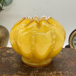 May include: A yellow glass decorative bowl with a round shape and a ruffled rim. The bowl has a textured surface with vertical beaded accents. The base is a darker shade of yellow. The bowl is sitting on a brown surface.