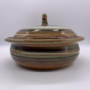 May include: A ceramic lidded serving dish with a handle. The dish features horizontal bands of brown, green, and tan colors. The lid has a small, curved handle with a swirl of colors. The dish has a wide rim.