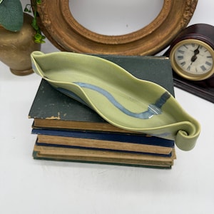 May include: A handmade, leaf-shaped ceramic dish in shades of green with a blue wave design. The dish has a curled end and sits atop a stack of vintage books. Decorative and functional, it could be used as a trinket tray or a small serving dish.