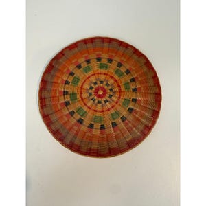 May include: A round, woven basket tray featuring a radial pattern of concentric circles. The design incorporates alternating bands of natural tan, deep red, forest green, and black fibers, radiating from a central star-shaped woven detail.