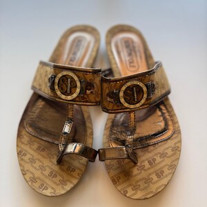 May include: A pair of bronze-toned thong sandals featuring a repeating chain link pattern and the letters SP printed on the footbed. Each sandal has a wide strap across the top with a circular metallic emblem containing the text Southpole.