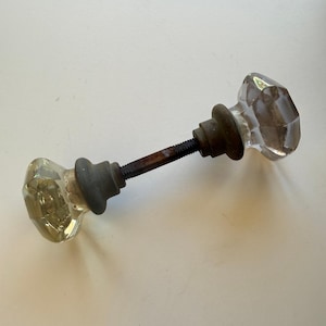 May include: A vintage door knob set featuring two clear, multifaceted glass knobs with octagonal shapes. The knobs are connected by a central metal spindle with a dark, weathered finish and visible rust, flanked by two circular metal base plates.