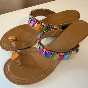 May include: A pair of tan leather sandals featuring a toe post and a wide strap adorned with multicolored beads and rectangular accents. The footbed is tan with the brand name Ann Marino embossed on the heel area of each sandal.