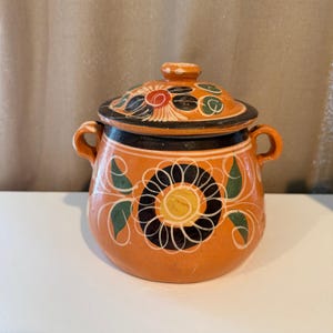 May include: A round terracotta pot with two side handles and a matching lid. The exterior features a hand-painted design with a large central black and yellow flower, green leaves, and white accents against an orange clay background.