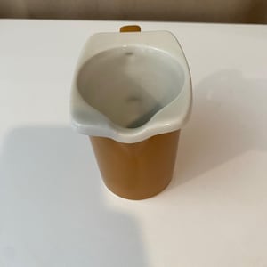 Może przedstawiać: Ceramiczny dzbanek na śmietankę o gładkiej, musztardowo-żółtej powierzchni zewnętrznej i kontrastującym białym wnętrzu. Naczynie posiada szeroką, zaokrągloną krawędź z wyraźnym dzióbkiem oraz mały uchwyt wystający z tylnej części pojemnika.