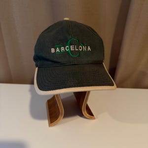 Könnte beinhalten: Dunkelgrüne Baseballkappe mit dem Wort "BARCELONA" in Weiß und Grün gestickt. Die Kappe hat einen gebogenen Schirm mit cremefarbenem Besatz. Der Hut wird auf einem Holzständer präsentiert.