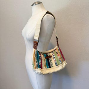 May include: A shoulder bag featuring a patchwork design of vertical fabric strips in various colors and patterns, including teal, orange, green, and cream. The bag has a cream-colored canvas strap and base, with brown leather-like accents.