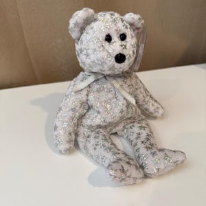 Puede incluir: Un oso de peluche blanco con patrones de estrellas plateadas y una cinta plateada alrededor del cuello. El oso tiene ojos negros y una nariz negra. Una pequeña etiqueta está adherida a la oreja.