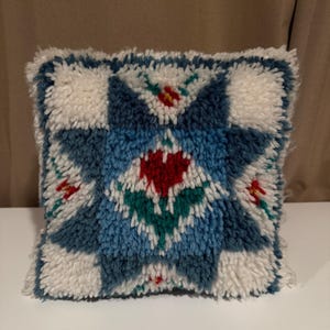 May include: Square decorative pillow with a geometric design. The pillow features a central starburst pattern in shades of blue, red, and green, surrounded by white and blue squares. The pillow has a textured, tufted appearance.