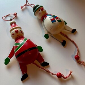 May include: Two wooden Christmas ornaments: a Santa Claus and a snowman. The Santa ornament is red with green and black accents, and the snowman is white with a colorful scarf. Both have dangling red string and are ready to hang.