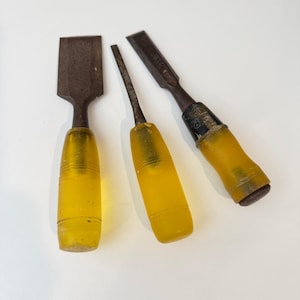 May include: Three vintage wood chisels with translucent yellow amber-colored plastic handles. Each tool features a metallic, oxidized steel blade of varying widths and shapes, showing signs of wear and age on the metal surfaces and handle ends.