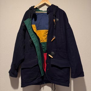 May include: A navy blue duffel coat with a hood and toggle closures. The coat has two front pockets and a colorful interior lining with green, yellow, blue, and red panels. The coat is hanging on a wooden hanger.