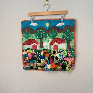 Peut inclure: Une tenture murale carrée en tissu représentant un paysage coloré avec deux maisons blanches, un lama blanc central, des arbres verts et un soleil jaune. La partie inférieure présente un arrangement dense de petites figurines en tissu.