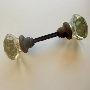 May include: A door knob set featuring two clear, faceted glass knobs with a hexagonal shape. The knobs are connected by a central threaded metal spindle and include dark, weathered metal bases that show signs of oxidation and age.