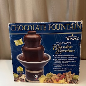 May include: A chocolate fountain in its original box. The box is dark blue with the words "CHOCOLATE FOUNTAIN" in gold lettering. The fountain is brown and has multiple tiers. The box also says "The Ultimate Chocolate Experience."
