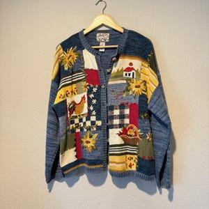 Könnte beinhalten: Eine langärmelige Strickjacke mit Knopfleiste und Patchwork-Design in Blau, Gelb, Rot und Weiß. Das Strickmuster zeigt gestickte Motive eines Hahns, eines kleinen Hauses, von Sonnenblumen und eines Obstkorbs auf einem blauen Hintergrund.
