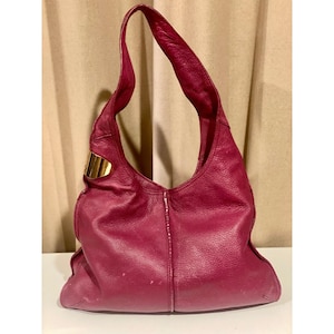May include: A magenta hobo-style shoulder bag crafted from textured leather. The bag features a single wide strap, a slouchy silhouette, and a prominent polished gold-tone metal accent plate attached to the side near the top handle attachment.
