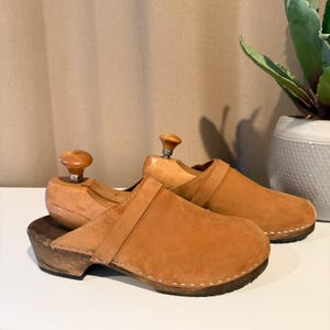May include: A pair of brown suede clogs with wooden soles and a strap across the top. The clogs are displayed with wooden shoe trees inside. The clogs are on a white surface.