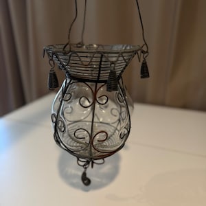 Puede incluir: Una linterna colgante de vidrio y metal. La linterna de vidrio transparente está encerrada en una jaula de metal decorativa con un asa de alambre para colgar. El metal tiene un acabado oscuro y envejecido. La linterna tiene forma bulbosa con espirales decorativas.