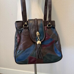 Può includere: Una borsa a spalla patchwork multicolore realizzata con pannelli in pelle nelle tonalità marrone scuro, blu navy e bordeaux intenso. La borsa presenta due tracolle, una patta centrale intrecciata con chiusura in metallo dorato e una nappa.