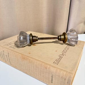 May include: A vintage-style door knob set featuring two clear, fluted glass knobs connected by a central brass-toned metal spindle. The hardware includes circular brass-toned bases at the neck of each glass knob, providing a classic, functional assembly for door hardware.