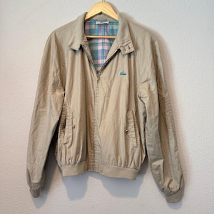 Puede incluir: Una chaqueta beige con cremallera, cuello, mangas largas con puños acanalados y dos bolsillos delanteros. El interior está forrado con una tela de cuadros azules y rosas, y un pequeño logotipo de cocodrilo verde está bordado en el pecho izquierdo.