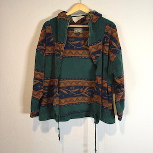 May include: A green fleece hooded jacket featuring a dark blue and orange geometric pattern with floral accents. The jacket has a full-length front zipper, long sleeves, and a drawstring hem with toggles. A small rectangular label is visible at the neck.