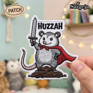 Puede incluir: Un parche bordado que muestra una zarigüeya gris de pie sobre un montículo de tierra marrón. La zarigüeya lleva una capa roja y sostiene una espada plateada hacia arriba. Sobre ella, la palabra HUZZAH está bordada en letras negras sobre fondo blanco.