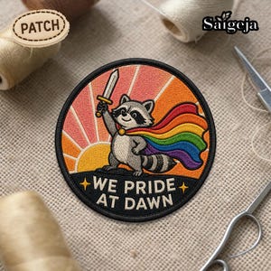 May include: Embroidered patch featuring a raccoon superhero with a rainbow cape and a sword, set against a sunset backdrop. The patch includes the text "WE PRIDE AT DAWN" in white lettering. The patch is round with a black border.