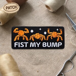 May include: Black rectangular patch with the text "FIST MY BUMP" in white. The patch features three orange crab-like figures flexing their arms. White stars are between the figures. The word "PATCH" is on a wooden tag.