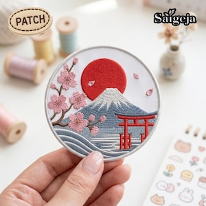 Peut inclure: Un écusson brodé circulaire sur fond blanc représentant un soleil rouge, une montagne bleue, des fleurs de cerisier roses sur une branche brune et un torii rouge. L'écusson a une bordure grise cousue et porte le texte PATCH en haut à gauche.