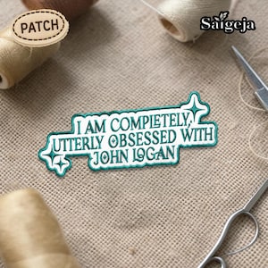 Puede incluir: Un parche rectangular con el texto "I AM COMPLETELY UTTERLY OBSESSED WITH JOHN LOGAN" en letras blancas, contorneado en turquesa. El parche está rodeado de diseños de estrellas turquesas. El fondo es una tela beige texturizada.