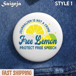 May include: A white button with the text "JOURNALISM IS NOT A CRIME" and "Free Lemon PROTECT FREE SPEECH" in blue and yellow. The button has a lemon graphic and the brand name "Saigeja" at the top left.
