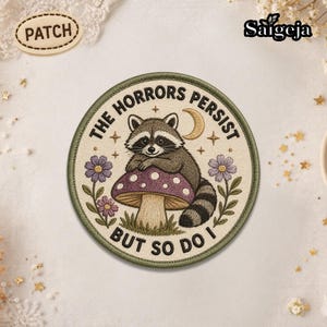 May include: Round patch with a raccoon sitting on a mushroom. The patch has the text "THE HORRORS PERSIST BUT SO DO I". The raccoon is gray and brown, the mushroom is purple and white, and the flowers are purple.