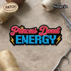 Può includere: Toppa ricamata con le parole "Princess Donut ENERGY" in rosa e blu, con una corona d'oro e fulmini. La toppa è nera con un bordo dorato. La parola "PATCH" è su una piccola etichetta di legno.