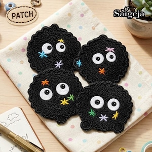 May include: Four circular black embroidered patches featuring large white eyes and small colourful star-shaped accents in yellow, blue, pink, orange, and green. Each patch has a scalloped edge and a fuzzy texture, arranged on a light fabric.
