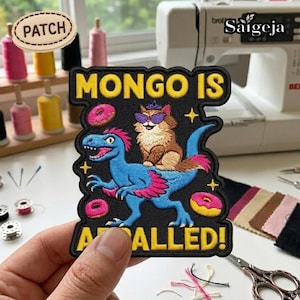 May include: Embroidered patch with the text "MONGO IS A CALLED!" featuring a blue dinosaur with pink accents and a cat wearing sunglasses. The patch includes donut and star designs, with a black background and yellow border.