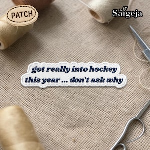 Puede incluir: Un parche bordado blanco con el texto azul marino "got really into hockey this year... don't ask why". El parche está sobre una tela beige, con carretes de hilo y unas tijeras en el fondo.