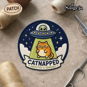 Puede incluir: Parche bordado circular que representa un gato de dibujos animados siendo abducido por un OVNI. El parche tiene un fondo azul marino, un rayo de luz verde y el texto "CATNAPPED - ABDUCTED & ADORABLE". El gato es naranja y blanco.