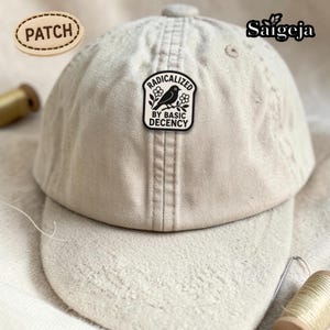 May include: A beige baseball cap with a curved brim. The cap features a black and white patch that reads "RADICALIZED BY BASIC DECENCY" with a bird and floral design. A wooden patch with the word "PATCH" is also visible.