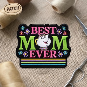 May include: Embroidered patch with the words "BEST MOM EVER" in pink and green, featuring a cartoon teapot character. The patch is black with colorful floral and striped accents. The word "PATCH" is on a separate wooden patch.