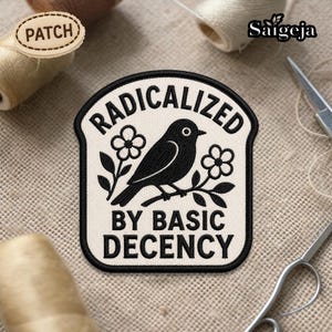 Puede incluir: Parche bordado crema con las palabras "RADICALIZED BY BASIC DECENCY" y un pájaro negro posado en una rama con flores. El parche tiene costuras negras. También es visible un pequeño parche de madera que dice "PATCH".